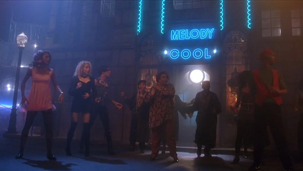 Graffiti Bridge (1990) movie still: wide — A city street at night features a club entrance glowing with blue neon letters. A small group of people…