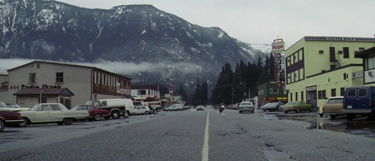First Blood (1982) movie still: establishing — A small town street lined with 1980s automobiles and low-rise commercial buildings. Large snow-capped…
