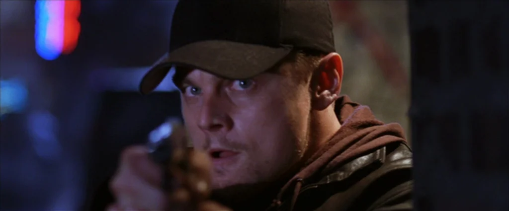 The Departed (2006) movie still: closeup — An urban setting at night with out-of-focus neon lights visible in the background. A man in a baseball…