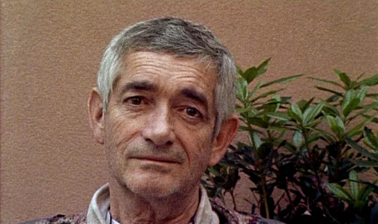 Jacquot de Nantes (1991) movie still: closeup — An older man stands outdoors with a textured brown wall behind him. A green leafy shrub is visible…