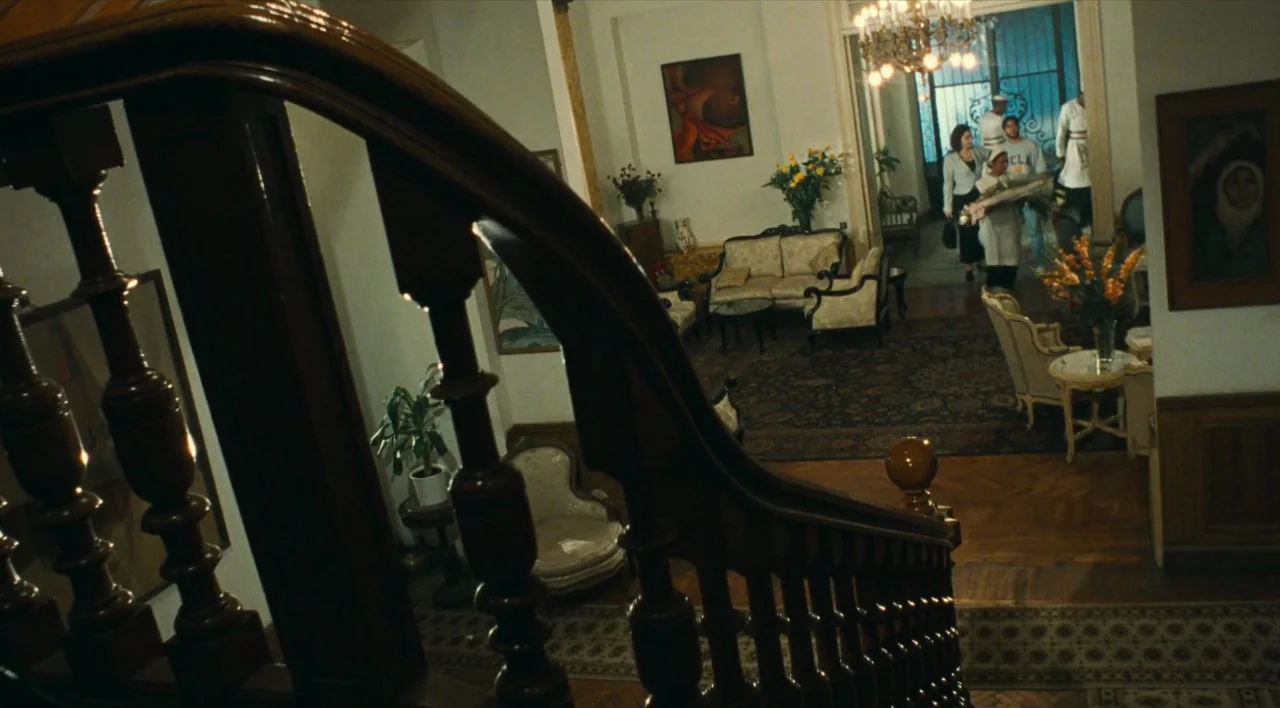 The Other Conquest (1999) movie still: wide — A spacious home entrance with a prominent wooden staircase and patterned area rugs. A group of four…