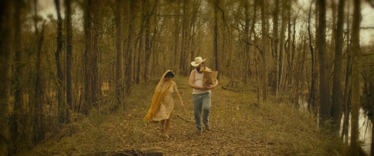 Blaze (2018) movie still: wide — A man and woman walk along a narrow, leaf-strewn dirt path in a swampy woodland. They move forward side by side…