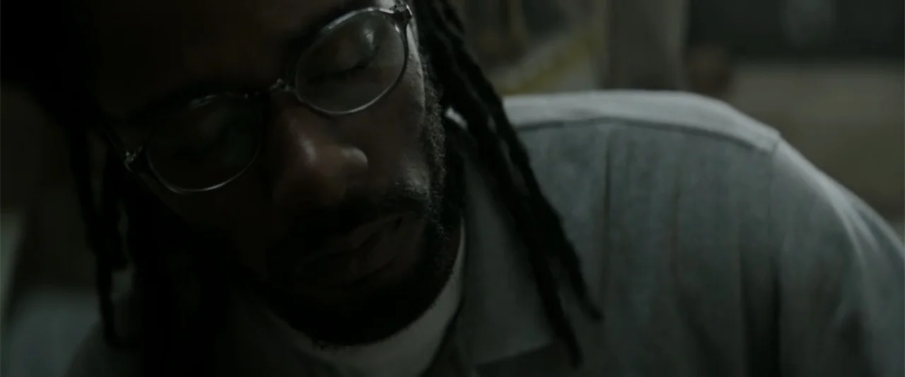 Crown Heights (2017) movie still: closeup — A dimly lit interior space with shadows cast across the subject. A Black man with locs and glasses sits…