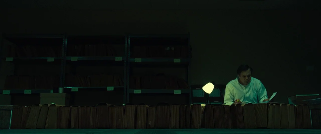 Dark Waters (2019) movie still: wide — A dark archives room is lined with metal shelving containing rows of brown files. A man sits at a desk under…