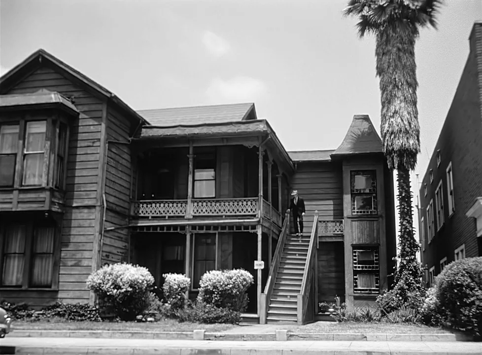 M (1951) (1951) movie still: wide — A two-story wood-sided Victorian house features a covered porch and an exterior staircase. A man in a suit…