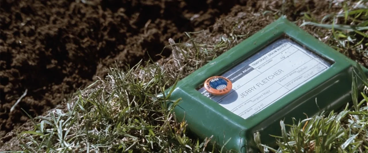 Conspiracy Theory (1997) movie still: closeup — A green plastic temporary grave marker for Jerry Fletcher sits in the grass. The name and details…