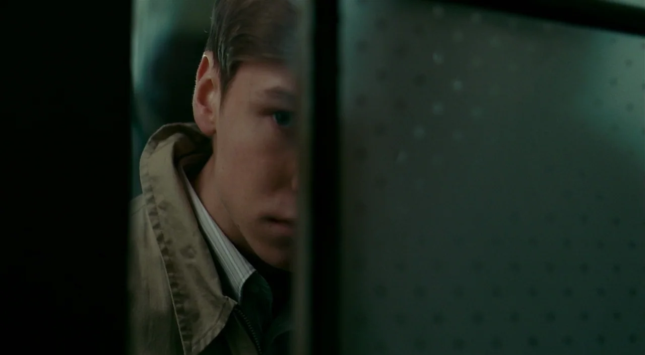 The Reader (2008) movie still: closeup — A dimly lit interior space features a glass partition with a subtle dot pattern. A teenage boy with light…