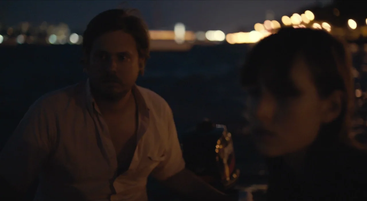 The Comedy (2012) movie still: medium — A man and a woman sit on a boat at night over dark water. Distant city lights create a bokeh effect against…