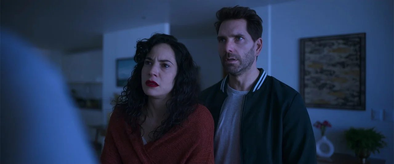 Evil Eye (2020) movie still: medium — A dimly lit room with blue ambient light cast on the subjects. A man and a woman stand together looking…