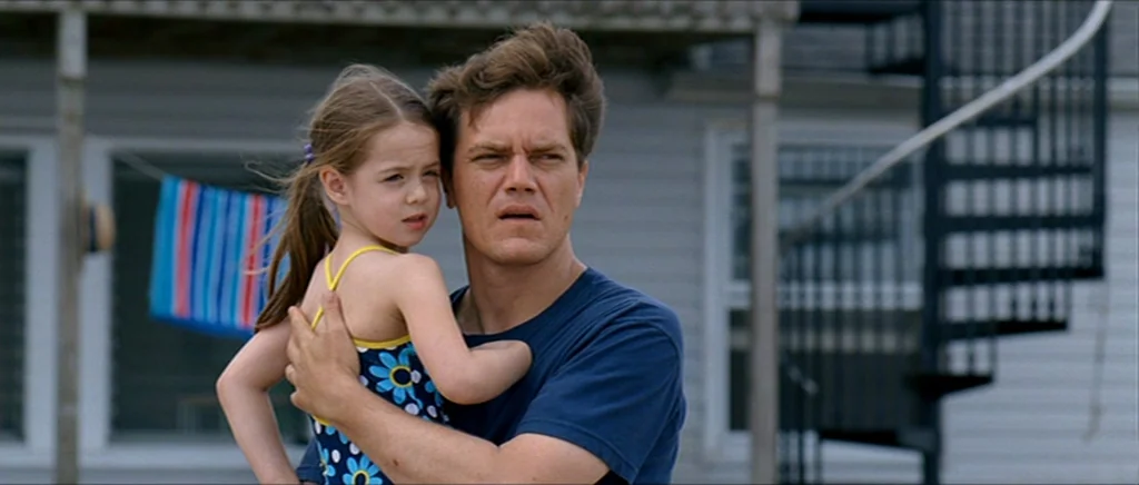 Take Shelter (2011) movie still: medium — A man holds a child in front of a house with a spiral staircase. A blue-striped towel hangs on a…