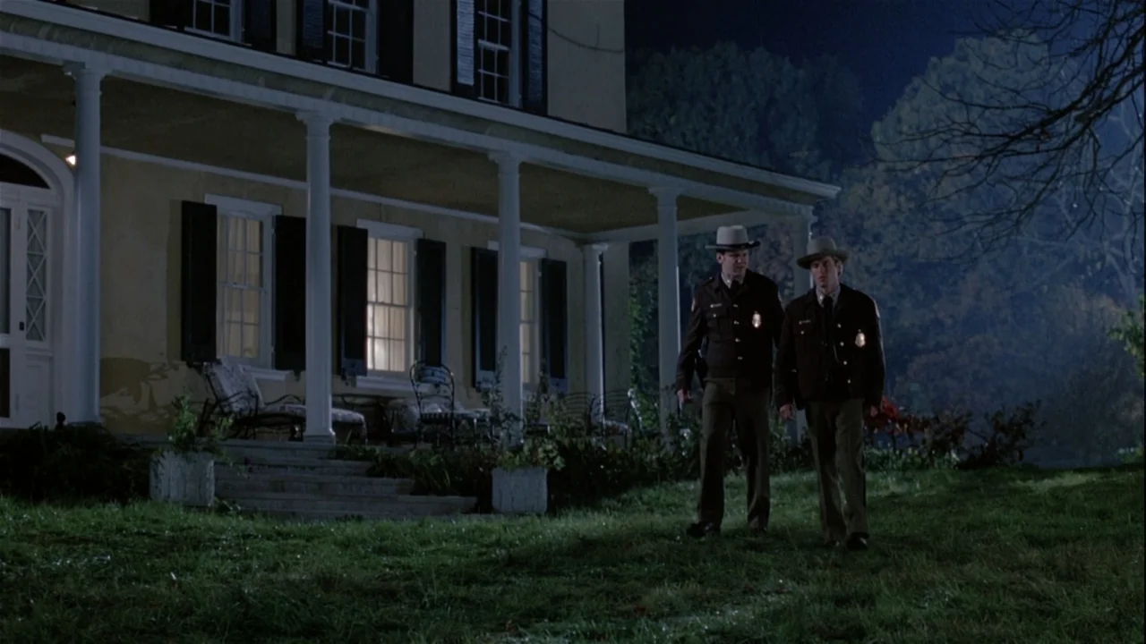 Boys (2021) movie still: wide — A historic yellow farmhouse features a wide porch with white columns and dark shutters. Two police officers in…