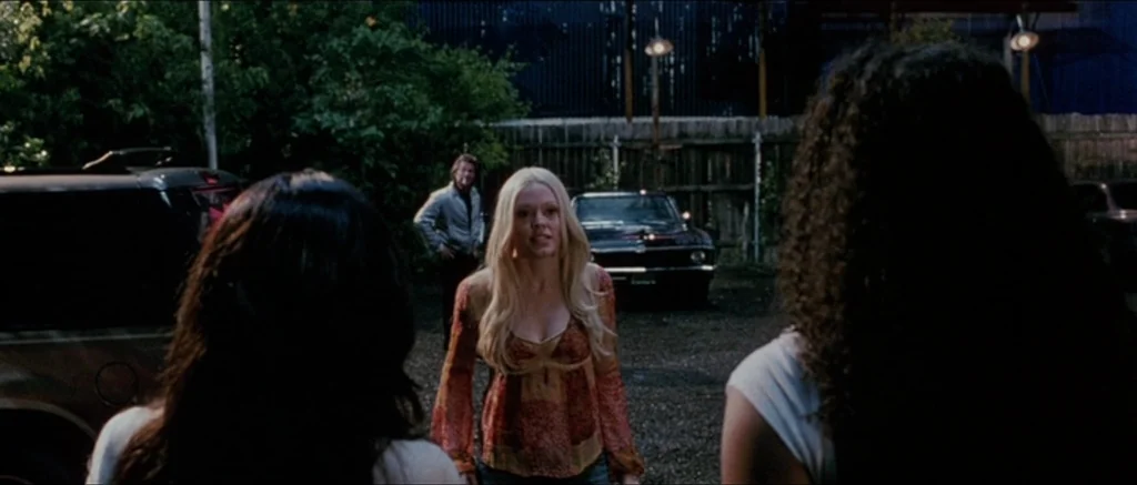 Death Proof movie still: medium — A gravel parking lot at night features a dark wooden fence in the back. A blonde woman stands center frame while…