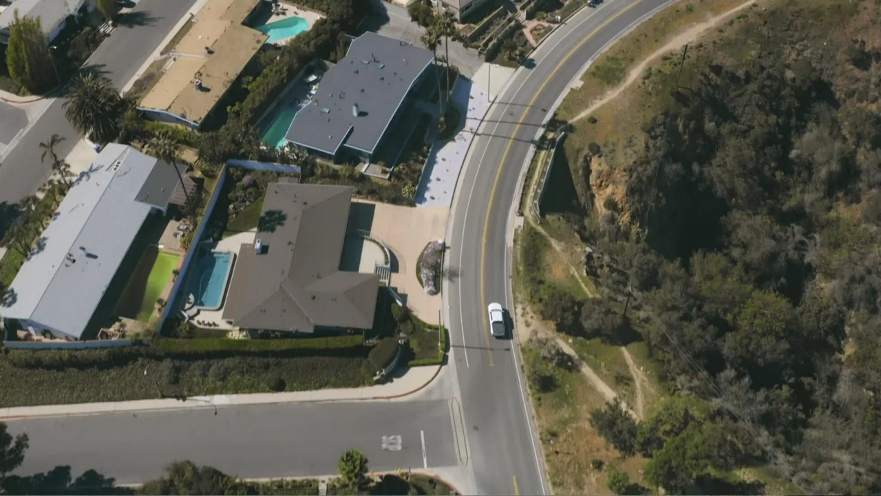 Introducing, Selma Blair (2021) movie still: establishing — An aerial view displays a winding road bordered by suburban houses with swimming pools.…