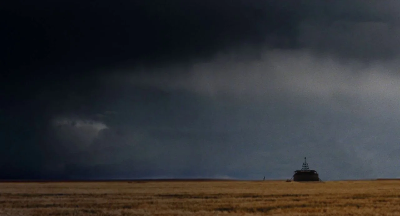 Tesla (2020) movie still: establishing — A vast, flat golden field sits beneath a massive, dark, threatening storm front. A small structure with an…