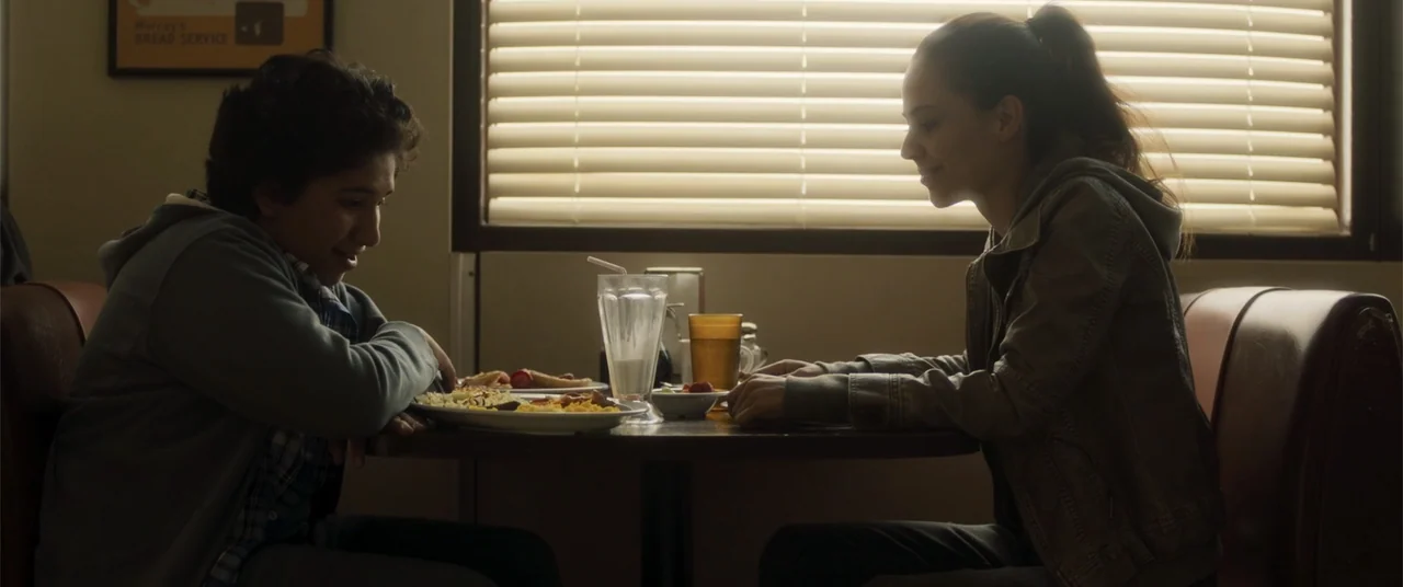 Phobias (2021) movie still: medium — A diner booth faces a large window with horizontal blinds letting in bright light. A teenage boy and a teenage…