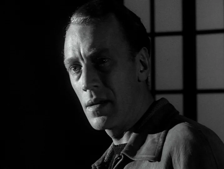 Hour of the Wolf (1968) movie still: closeup — A man stands in a dark room with a window grid background. The man gazes off-screen while the rest of…