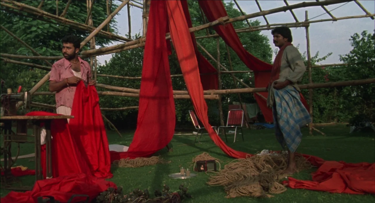 Monsoon Wedding (2001) movie still: fullbody — A grassy outdoor area features a skeletal bamboo frame hung with long red cloth. Two men stand within…