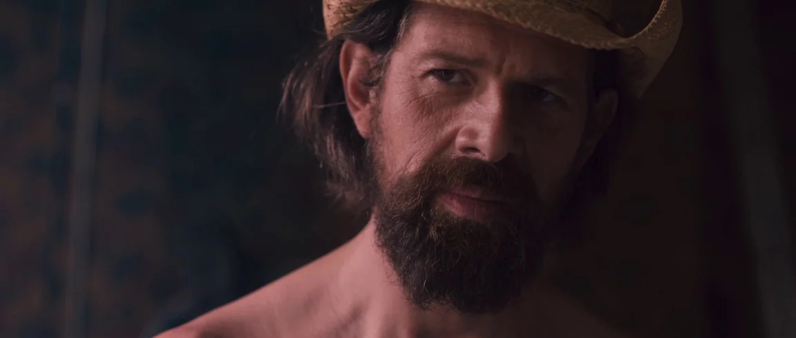 Broken Circle Breakdown (2012) movie still: closeup — A dimly lit indoor space with a dark, indistinct backdrop behind the subject. A shirtless man…