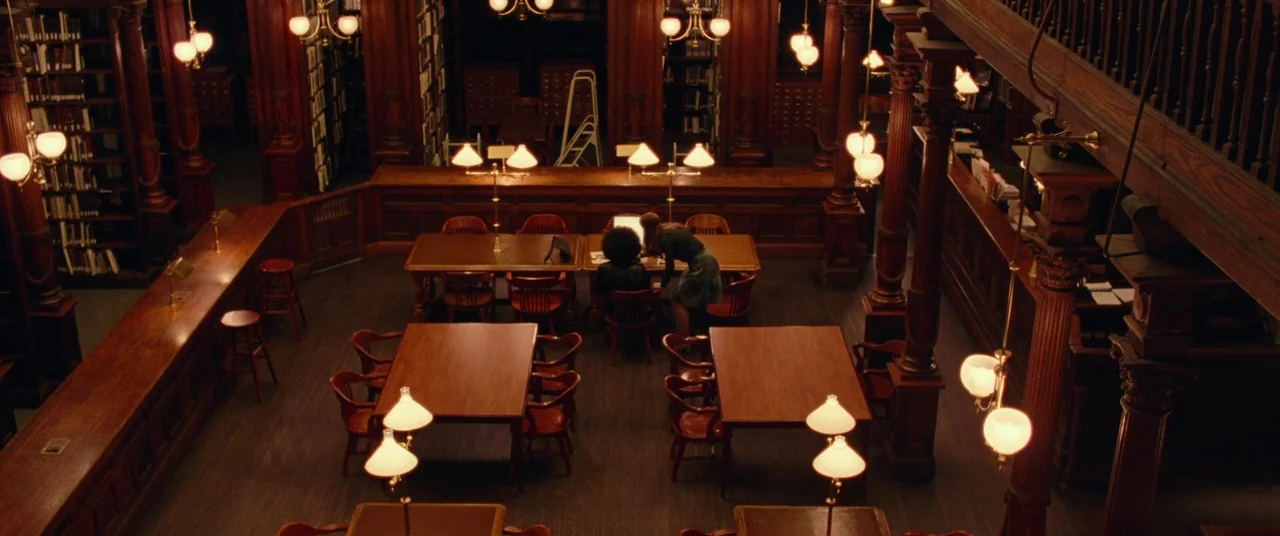 BlacKkKlansman (2018) movie still: wide — A spacious library interior features dark wood bookshelves and rows of study tables. Two women sit at a…