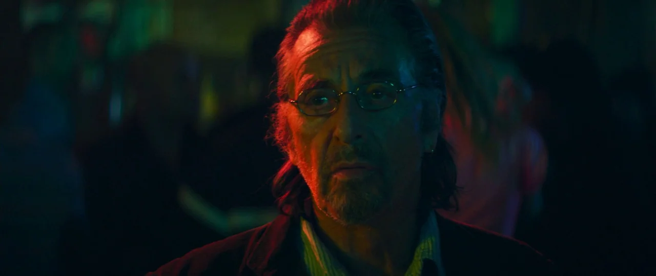 Manglehorn (2014) movie still: closeup — A dimly lit space with vibrant red and green neon lighting illuminating the subject. An older man stands in…
