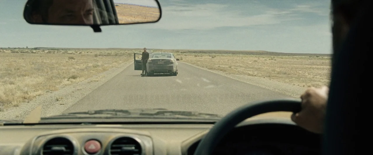 The Rover (2014) movie still: wide — An arid, flat landscape stretches out along a paved road under a bright sky. A man stands outside his…