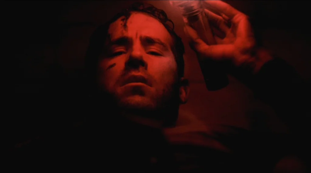Buried (2010) movie still: closeup — A man lies on his back inside a confined, dark wooden space. He holds a single red glowing light source near…