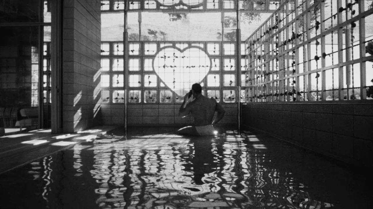 Divinity (2023) movie still: wide — A tiled bath area features a large window with a heart-shaped cutout design. A muscular man sits in the shallow…