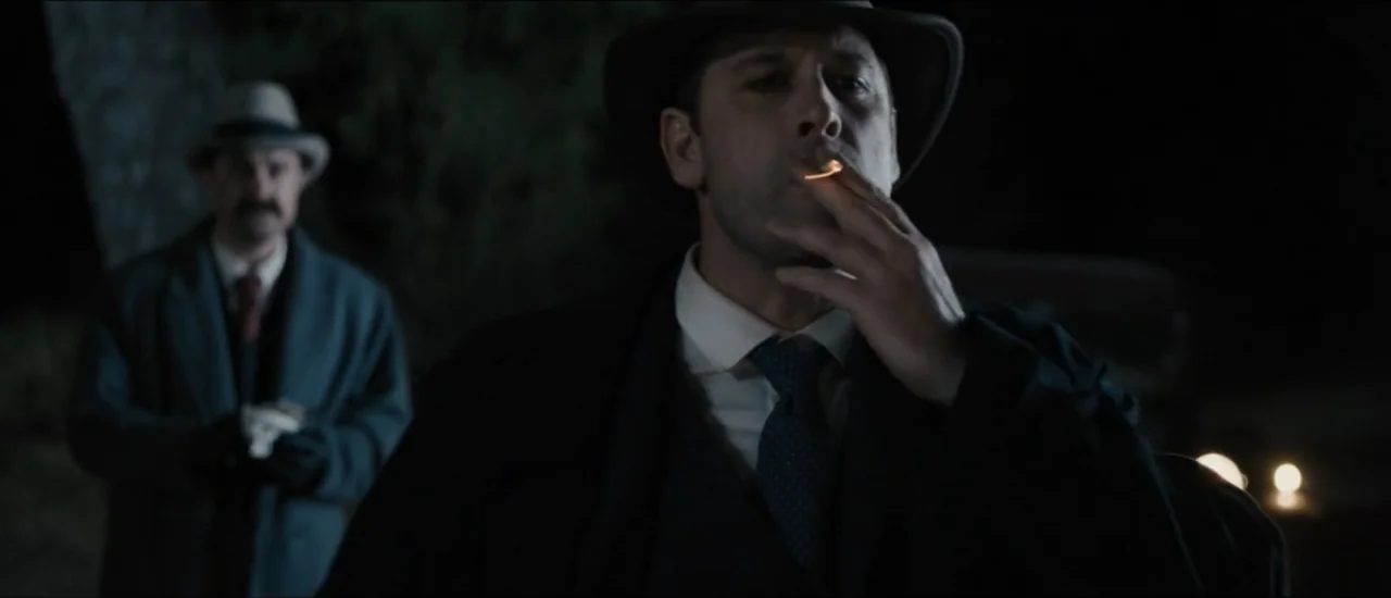 Fear Below (2025) movie still: medium — A dark outdoor area at night with distant car headlights providing low light. A man in a suit smokes in the…