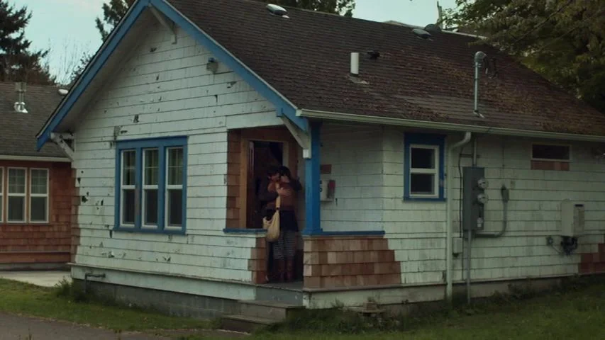 Touchy Feely (2013) movie still: wide — A weathered white cottage with blue trim features a covered front porch and entry. Two people stand closely…