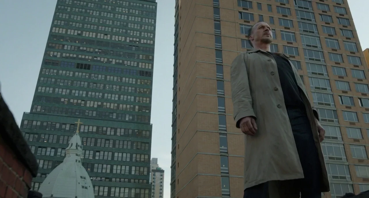 Birdman (2014) movie still: fullbody — A rooftop terrace provides a view of tall city apartment buildings and a church. A man in a beige coat stands…