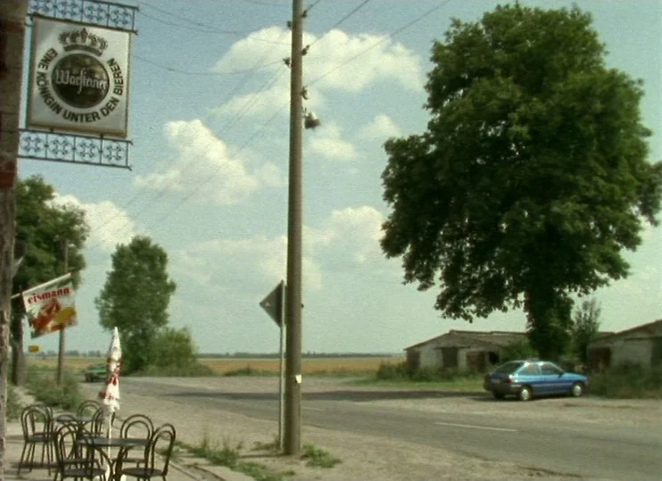 From The East (1993) movie still: wide — A rural roadside scene features an outdoor cafe seating area and a single car. A Warsteiner beer sign hangs…