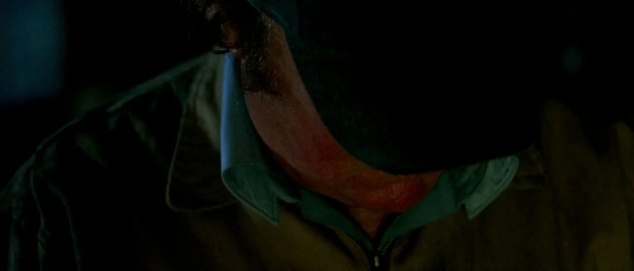 Haute Tension (2003) movie still: closeup — A dark indoor setting with limited illumination on the subject. A man wearing a cap and jacket looks…