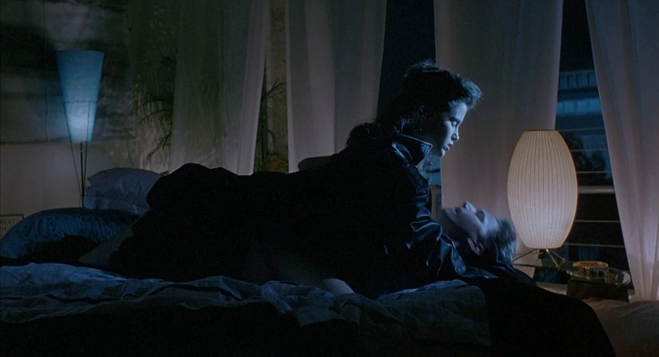 Less Than Zero (1987) movie still: medium — A dark bedroom with white curtains and modern lamps framing the central figures. A woman in a black…