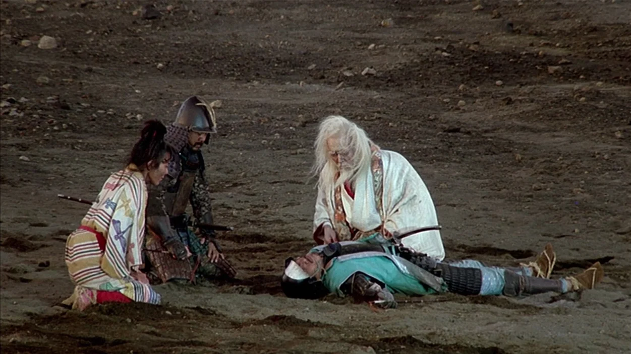 Ran (1985) movie still: wide — A group of people kneels on a vast expanse of barren, sandy, rocky earth. An elderly man in white robes examines a…