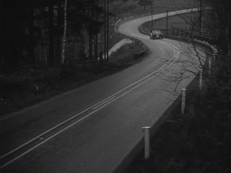 A Lesson In Love (1954) movie still: establishing — A winding asphalt road curves through a dense forest with tall trees and guardrails. A small…