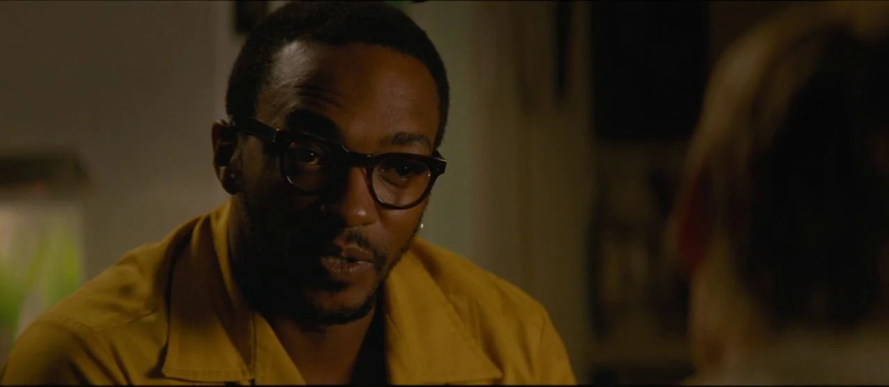 Seberg (2019) movie still: closeup — An interior space is dimly lit with deep shadows surrounding the subjects. A man in a yellow shirt looks toward…