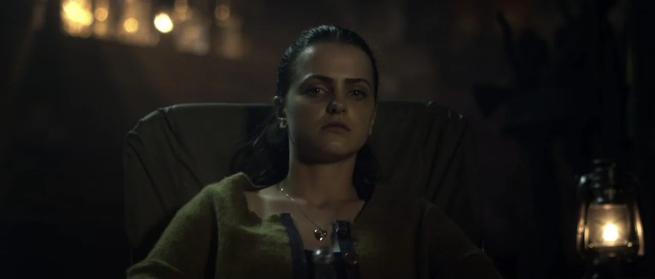 The Lullaby (2019) movie still: medium — A woman sits in a chair in a dark, indistinct interior space illuminated by a lantern. The woman looks…