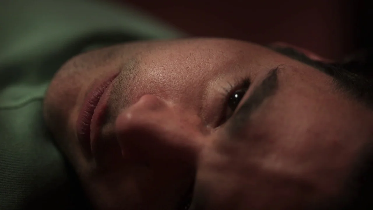 Piercing (2018) movie still: closeup — The frame shows a close-up of a man's face resting against a surface. He lies on his side with his eyes open…