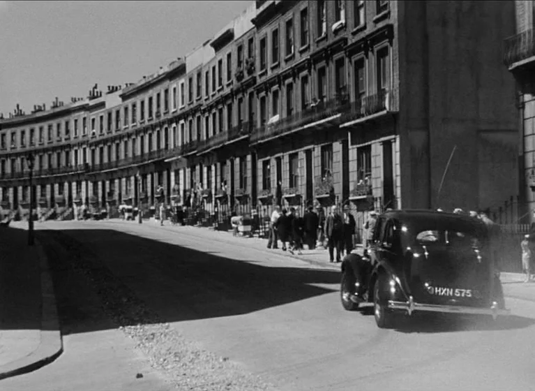 The Blue Lamp (1950) movie still: wide — A long, curved row of Victorian-style terraced houses lines a quiet city street. A black vintage automobile…