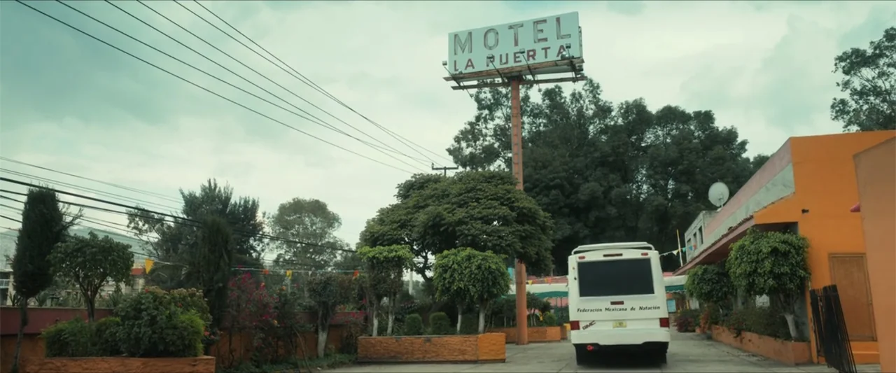Dive (2013) movie still: establishing — A motel courtyard features a prominent pole-mounted sign and a parked white minibus. Landscaped greenery and…