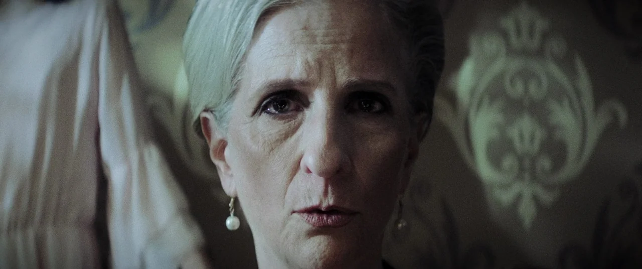 La Llorona (2019) movie still: closeup — An older woman is framed in a close-up against a dark, ornate patterned wall. She gazes directly into the…