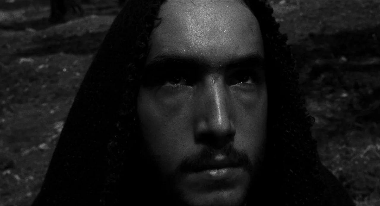 The Gospel According To St. Matthew (1964) movie still: closeup — A person in a hood stands in a dark, textured outdoor environment. The individual…