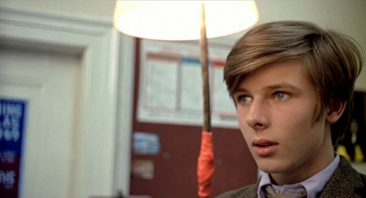 Deep End (1970) movie still: closeup — A school setting features a cork board with colorful charts in the background. A young man stands in the…