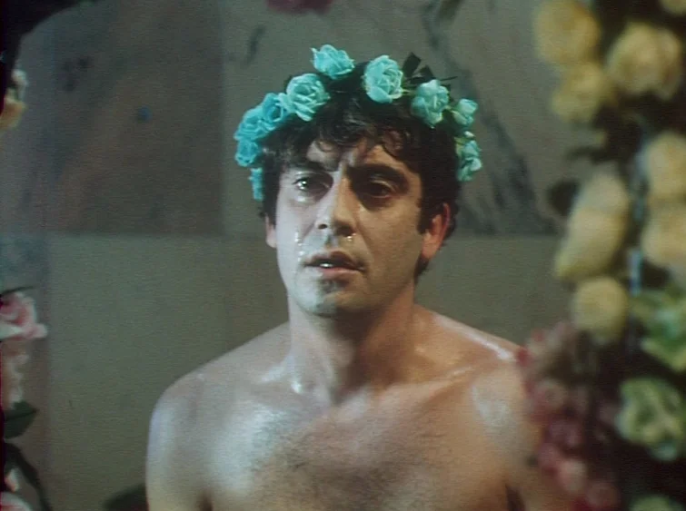 Hermitage (1968) movie still: medium — A man stands indoors surrounded by thick vertical arrangements of soft, pale-colored flowers. He has a crown…
