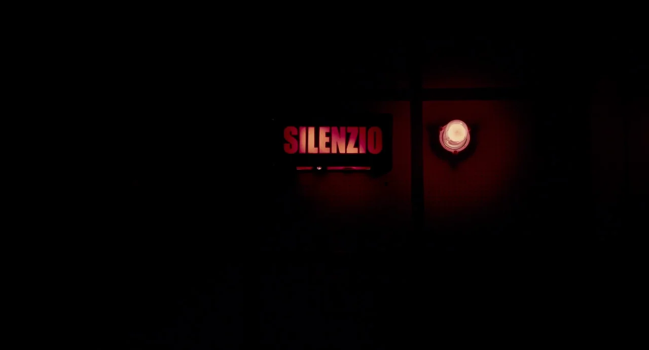 Berberian Sound Studio (2012) movie still: wide — A dimly lit wall features a glowing red sign and a single circular lamp. The word SILENZIO is…
