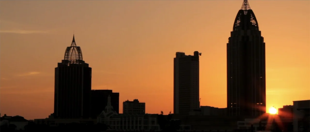 Rage (2014) movie still: wide — A metropolitan skyline is silhouetted against a vibrant orange and yellow sunset horizon. Tall skyscrapers dominate…