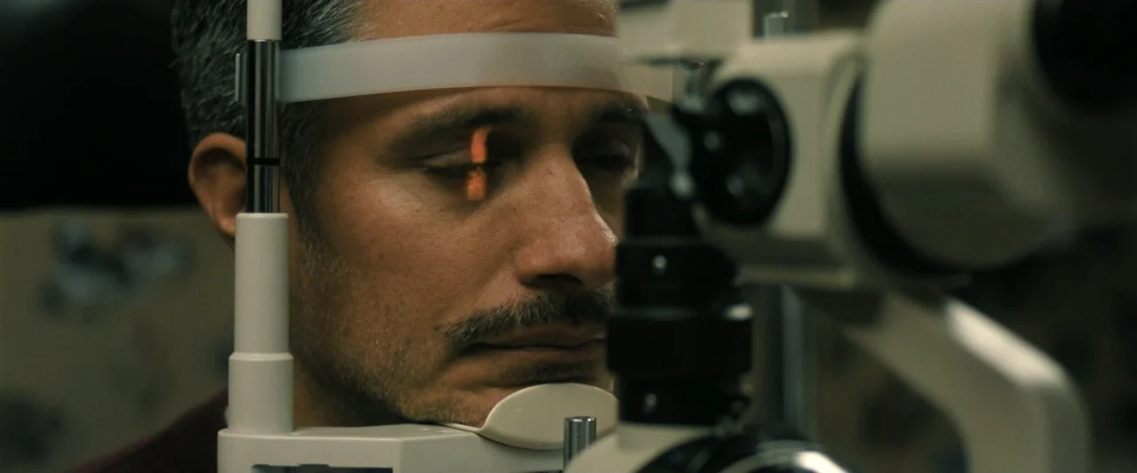 Holland (2025) movie still: closeup — A clinical interior space contains an ophthalmic slit lamp instrument with a chin rest. A man rests his head…
