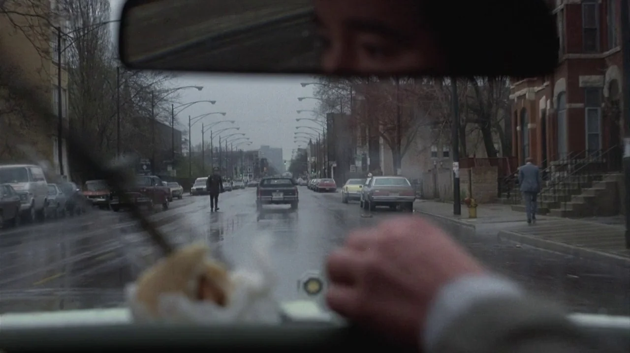 Thief (1981) movie still: wide — A wet, overcast urban street is lined with parked cars and brick row houses. A police officer stands in the middle…