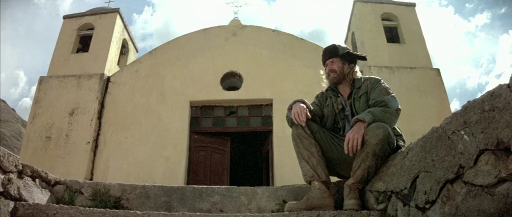 The Big Blue (1988) movie still: fullbody — A stucco church with two bell towers stands against a cloudy, bright sky. A bearded man sits on the…