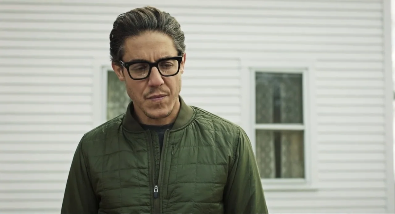 Salvation (2025) movie still: medium — A man stands before the side of a plain white horizontal-sided house. The man wears an olive green quilted…